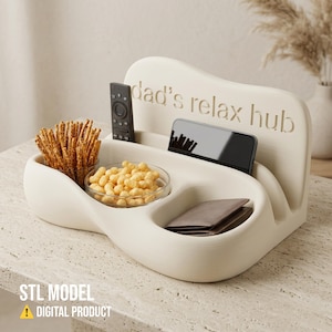 Puede incluir: Una bandeja organizadora curva de color crema con el texto en relieve "dad's relax hub". La bandeja incluye varios compartimentos que contienen un mando a distancia negro, un smartphone, una cartera marrón y dos cuencos de cristal con aperitivos.