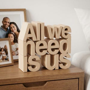 May include: A wooden block sculpture spelling "All we need is us" in a stacked arrangement. The letters are a light beige color and sit on a wooden nightstand. Two framed family photos are in the background.