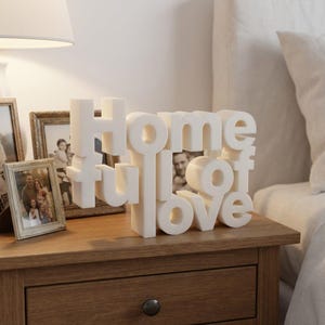 K&ouml;nnte beinhalten: Eine wei&szlig;e 3D-Skulptur mit der Aufschrift "Home full of love" steht auf einem Nachttisch aus Holz. Die Skulptur befindet sich vor gerahmten Familienfotos. Eine wei&szlig;e Lampe und ein Bett mit wei&szlig;er Bettw&auml;sche sind im Hintergrund.