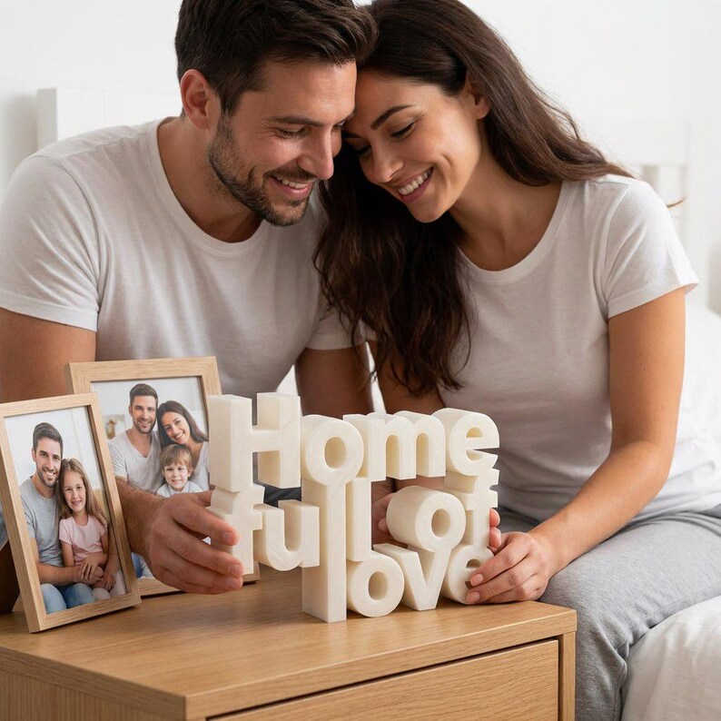 K&ouml;nnte beinhalten: Auf einem Nachttisch aus Holz stehen zwei gerahmte Familienfotos und eine wei&szlig;e, dreidimensionale Skulptur mit der Aufschrift "Home full of love". Ein Paar l&auml;chelt und betrachtet die Skulptur.