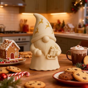 Op de afbeelding: Een crèmekleurig kabouterbeeldje met een hoge hoed versierd met gouden rendieren. De kabouter houdt een cadeau op een slee vast. De scène omvat een peperkoekhuisje, koekjes en een mok warme chocolademelk, wat een feestelijke sfeer creëert.