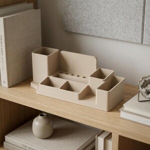 May include: A beige desk organizer with multiple compartments, including pen holders and small trays. The organizer sits on a wooden shelf with books and a small vase. The design is minimalist and functional, suitable for office or home use.
