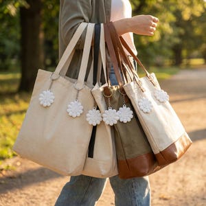 May include: A collection of canvas tote bags in various colors, including beige, olive green, and black. Each bag features a matching strap and a white daisy-shaped charm. The bags are carried by a person walking outdoors.