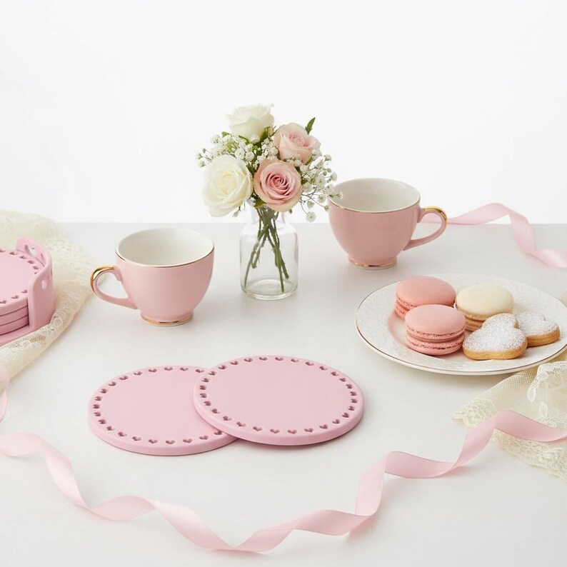 May include: A still-life arrangement featuring pink teacups with gold rims, coasters, and a small vase of flowers. Macarons and cookies are on a white plate. Pink ribbons and lace add to the delicate aesthetic. The scene is set on a white surface.
