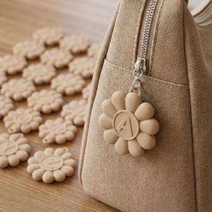 May include: A beige canvas bag with a silver zipper and a flower-shaped charm. The charm has the letter "A" in the center. Scattered around the bag are several beige flower-shaped coasters.