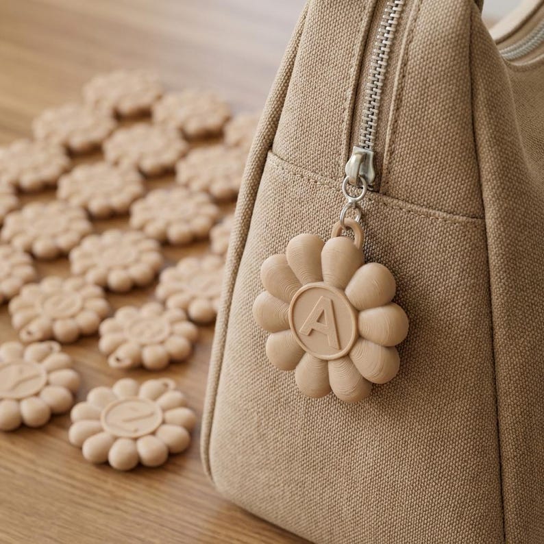 May include: A beige canvas bag with a silver zipper and a flower-shaped charm. The charm has the letter "A" in the center. Scattered around the bag are several beige flower-shaped coasters.