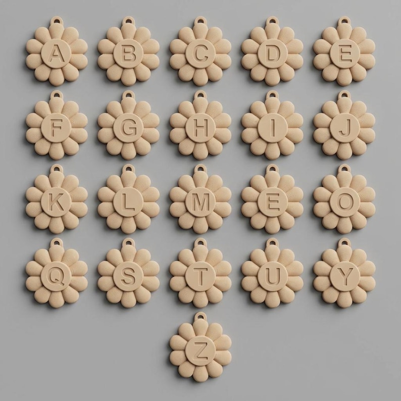 May include: A collection of beige daisy-shaped charms, each with a different letter of the alphabet in the center. The charms are arranged in rows and columns against a gray background. Each charm has a small loop at the top.