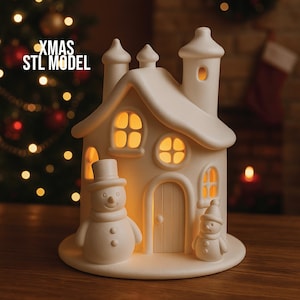 May include: A white ceramic Christmas house with a snowman and a smaller snowman figurine. The house has lit windows and a chimney. The text "XMAS STL MODEL" is visible on the left side of the image. The house is on a wooden surface.