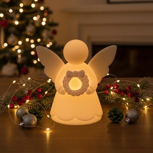 May include: A glowing angel-shaped Christmas light with outstretched wings and a wreath. The angel is holding a wreath with a bow. The light is surrounded by Christmas decorations, including a Christmas tree, pine needles, and ornaments.