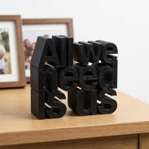 May include: Black 3D-printed decorative block with the phrase "All we need is us". The letters are in a bold, sans-serif font. The block is displayed on a wooden surface, with framed photos in the background.