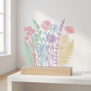 Op de afbeelding: Een pastelkleurige bloemensculptuur met verschillende bloem- en bladontwerpen. De bloemen zijn in roze, blauw, paars en geel, gemonteerd op een rechthoekige houten basis. Een decoratief item.