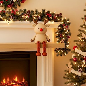 May include: A beige reindeer figurine with brown antlers, arms, and legs, and a red nose. The reindeer is perched on a wooden mantelpiece decorated with Christmas garland and lights. A Christmas tree is in the background.