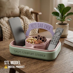 以下が含まれることがあります： 「DAD'S COMFORT CORNER」と「BEST DAD EVER」の文字が入ったツートーンのトレイ。トレイにはスマートフォン、リモコン、ナッツとプレッツェルが入ったスナックボウルが入っています。デジタル製品です。