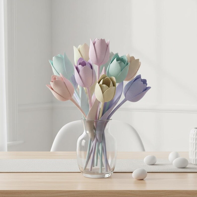 May include: A clear glass vase holds a bouquet of pastel-colored tulips. The tulips are in shades of pink, purple, green, and yellow. The vase sits on a wooden table with a white runner. White eggs are scattered on the table.