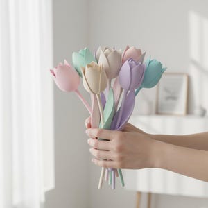 May include: A bouquet of pastel-colored tulip flowers in shades of pink, cream, lavender, and mint green. The tulips are held in a person's hands, with the stems and leaves visible. The flowers are a soft, matte material.