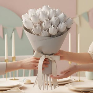 May include: A bouquet of white tulips wrapped in gray paper and tied with a white ribbon. The bouquet is being held by two people, with candles and plates on a table in the background.