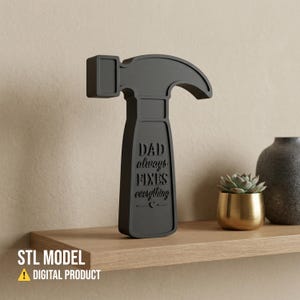 Könnte beinhalten: Ein dunkelgraues, hammerförmiges Dekorationsobjekt mit dem Text "DAD always FIXES everything" auf dem Griff. Es steht auf einem Holzregal mit einer kleinen Sukkulente in einem goldenen Topf und einer grauen Vase. Digitales Produkt.