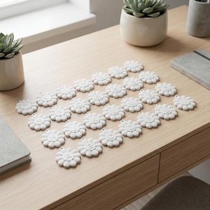 May include: A collection of white, daisy-shaped charms with engraved numbers and smiling faces arranged on a light wood surface. The charms are evenly spaced, suggesting a decorative or organizational purpose. Small potted succulents and a notebook are also visible.