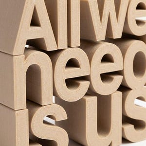 May include: Close-up of stacked, beige 3D-printed letters spelling "All We Need Is Us." The letters are blocky and have a textured surface. The composition is a close-up, emphasizing the message and the tactile quality of the letters.
