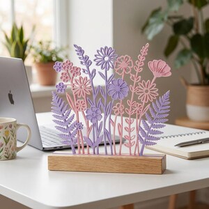 Op de afbeelding: Een decoratief bloemstuk met lasergesneden bloemen in roze en paarse tinten, gemonteerd op een natuurlijke houten basis. Het ontwerp omvat verschillende bloemsoorten en bladeren, wat een lenteachtige uitstraling cre&euml;ert. Het stuk staat op een witte bureau.