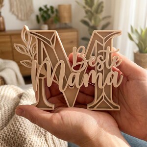 Op de afbeelding: Een beige houten decoratief stuk in de vorm van de letter "M" met de woorden "Beste Mama" in script. De letter is gelaagd met een bladontwerp aan de linkerkant. Het stuk wordt in de handen van een persoon gehouden.
