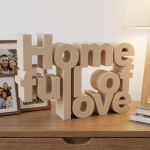 K&ouml;nnte beinhalten: Eine beige Holzskulptur schreibt "Home full of love" in einem gestapelten, dreidimensionalen Design. Die Skulptur steht auf einer Holzoberfl&auml;che, mit gerahmten Fotos und einer Lampe im Hintergrund. Die Buchstaben sind in einer serifenlosen Schriftart.