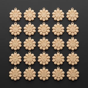 May include: A collection of beige flower-shaped pendants, each with a different letter of the alphabet in the center. The pendants have a small loop at the top for hanging. The background is a solid black.