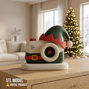 May include: A cream and dark green camera-shaped object featuring a red elf hat with a white pom-pom, a red belt with a yellow buckle, and the text "ELF CAM" printed on the front, all resting on a cream-coloured rectangular base.