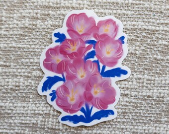 May include: A sticker depicting a cluster of pink flowers with yellow centres and blue leaves. The flowers are painted with visible brushstrokes, and the sticker has a white border. The background is a textured, neutral-coloured surface.