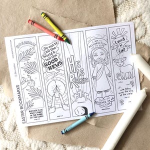 May include: A white sheet of paper featuring five vertical colouring bookmarks with religious illustrations and text including "HOSANNA!", "HE IS RISEN", and "LAMB OF GOD". Three wax crayons in red, yellow, and blue are placed on the paper.