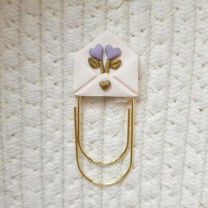 May include: A gold-toned metal paperclip topped with a pale pink envelope-shaped ornament. The envelope features two small lavender heart-shaped flowers with gold stems and leaves, and a single small gold heart centred below the flowers.