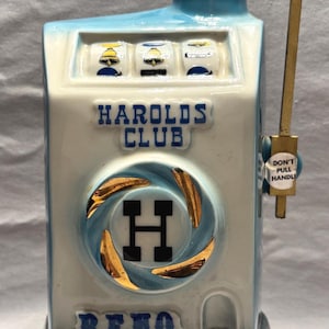 May include: A ceramic decanter shaped like a slot machine from Harolds Club in Reno. The white and light blue body features a gold letter H, a side lever with a label reading Don't Pull Handle, and a base label for Beam Kentucky Straight Bourbon Whiskey.
