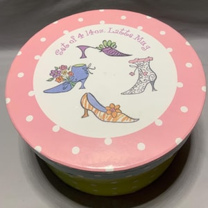 May include: A round container lid with a pink background and white polka dots. The center features the text Set of 4 14oz. Latte Mug and illustrations of four different shoes, including a blue shoe with flowers, a purple heel, a patterned boot, and an orange striped shoe.