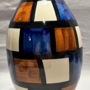 May include: A ceramic vase with a geometric design. The vase features a dark blue rim and base, with a body divided into sections of white, brown, orange, and blue, outlined in black. The vase is approximately 10 inches tall.