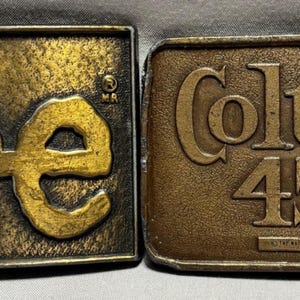 May include: Two rectangular brass belt buckles. The left buckle features the word Lee in raised lettering. The right buckle displays the text Colt 45 Malt Liquor by National with a horseshoe and figure graphic, all in a raised metallic finish.