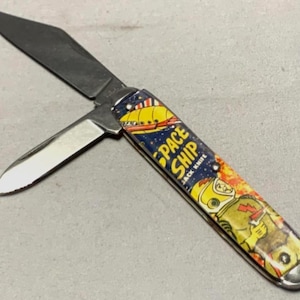 May include: A pocket knife with two blades, one open and one closed. The handle features a colorful design with the words "SPACE SHIP JACK KNIFE" and an illustration of a spaceship and an astronaut in a yellow space suit.