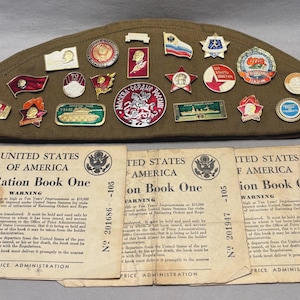 May include: An olive drab military cap adorned with numerous vintage pins and badges. Below, three aged "War Ration Book One" booklets from the United States of America are displayed, featuring text and official seals.