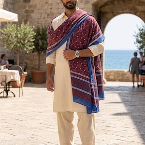 May include: A beige long-sleeved tunic and matching trousers paired with a maroon and blue patterned shawl. The shawl features a repeating circular motif and a solid blue border. The outfit is completed with brown leather sandals and a silver watch.