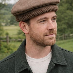 May include: A brown wool hat with a rounded crown and a textured band. The hat is worn by a person wearing a dark green jacket and a white t-shirt. The background features a blurred outdoor scene.
