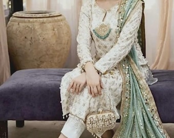 Könnte beinhalten: Eine weiße langärmelige Tunika und eine passende Hose mit aufwendiger Silberstickerei und Schlüsselloch-Ausschnitt. Ein hellgrüner Dupatta mit Goldbesatz und Spiegelarbeiten liegt über der Schulter. Eine kleine, rechteckige, goldfarbene Clutch mit Blumenverzierungen wird auf dem Schoß gehalten.