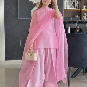 May include: A light pink sleeveless tunic paired with matching wide-leg trousers and a sheer organza dupatta. The outfit is accessorized with a small beige structured handbag featuring gold hardware and silver embellished open-toe slide sandals.