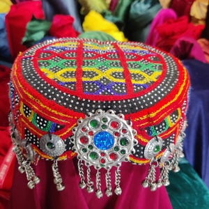 May include: A colorful, embroidered cap with a round crown. The cap features a red base with intricate patterns in yellow, green, and blue. Silver embellishments and dangling charms adorn the front. The cap is displayed on a deep red surface.