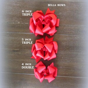 Pu&ograve; includere: Sono mostrati tre fiocchi rossi per capelli di diverse dimensioni. Il pi&ugrave; grande &egrave; un fiocco triplo da 15 cm, seguito da un fiocco triplo da 13 cm e un fiocco doppio da 10 cm. Il testo "BELLA BOWS" &egrave; visibile.