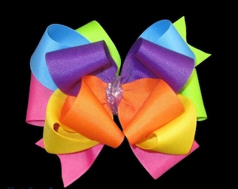 May include: A rainbow-colored hair bow with a glittery center. The bow is made of multiple layers of ribbon in the colors of the rainbow, including pink, yellow, orange, green, blue, and purple.
