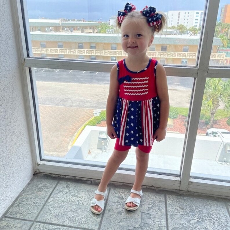 May include: A young girl wearing a red, white, and blue sleeveless dress with stars and stripes. She is wearing white sandals and has her hair pulled back with red, white, and blue bows.