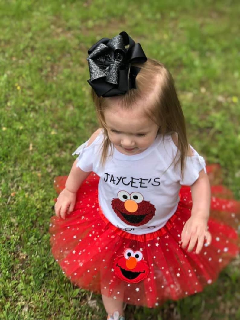 Może przedstawiać: Str&oacute;j dla dziecka składający się z białej koszulki z napisem "JAYCEE'S" i motywem Elmo, w połączeniu z czerwoną sp&oacute;dniczką tutu ozdobioną białymi gwiazdkami i aplikacją Elmo. Duża czarna kokarda zdobi głowę dziecka.