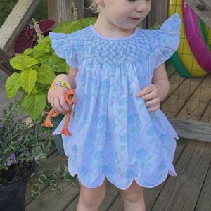 May include: A light blue dress with a smocked bodice, flutter sleeves, and a scalloped hem featuring a floral pattern. The dress is paired with a large light blue hair bow and white sandals. The garment has a light pink trim along the neckline and hem.