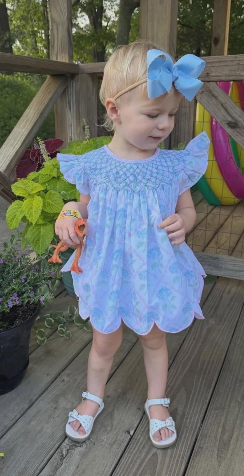 May include: A light blue dress with a smocked bodice, flutter sleeves, and a scalloped hem featuring a floral pattern. The dress is paired with a large light blue hair bow and white sandals. The garment has a light pink trim along the neckline and hem.