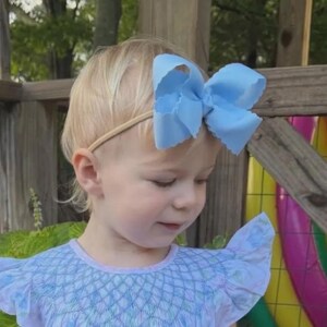 May include: A light blue hair bow with scalloped edges attached to a thin, neutral-colored elastic headband. The bow is large and features a structured, multi-layered design, providing a prominent accessory for the hair.