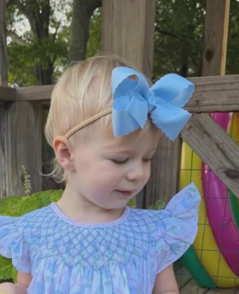 May include: A light blue hair bow with scalloped edges attached to a thin, neutral-colored elastic headband. The bow is large and features a structured, multi-layered design, providing a prominent accessory for the hair.